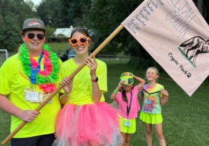 Augusta 4-H camp counselors prepare for a teambuilding activity at Junior 4-H Camp. Two camp counselors dressed in brightly colored, whimsical clothes and sunglasses hold a flag for their "pack" with two campers, also brightly dressed, standing in the background.